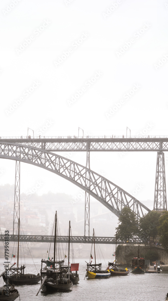 Wooden rabelos boats docked on the Douro River in Porto with wine barrels inside and the Don Luis I steel bridge with tourists strolling with umbrellas shrouded in fog on a gray rainy day.