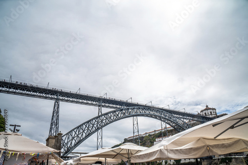 Canvas roofs of the traditional Ribeira market, on the Vila de Gaia promenade in Porto, with the Dom Luis I bridge in the background with pedestrians walking under a cloudy sky. © sirbouman
