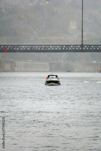 Boat sailing below the lower platform of the Don Luis steel bridge, with people walking with umbrellas due to the light rain and fog that surrounds the environment.