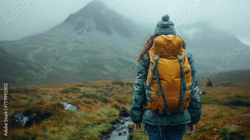  Person with backpack hikes in open field before towering mountain shrouded in fog