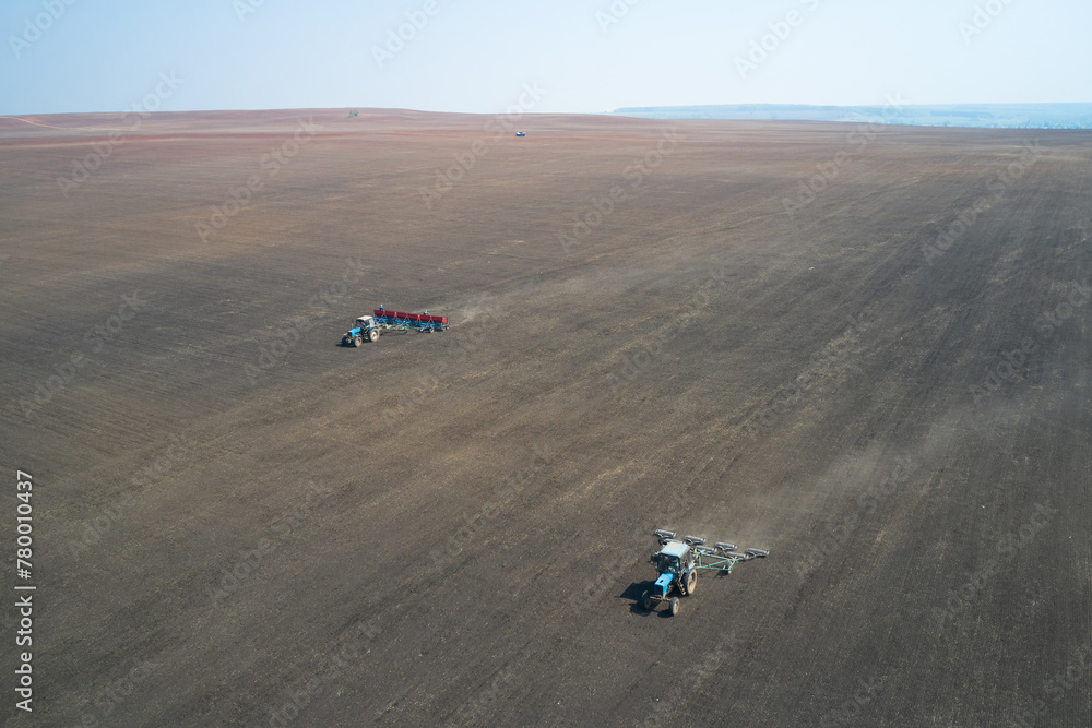 Sowing campaign. Sowing wheat in the field. Behind the tractor with a seeder works tractor with rollers. Shooting from a drone. - obrazy, fototapety, plakaty 