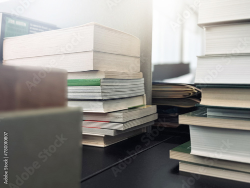 Stacks of books are on an office desk in blurred focus, illuminated by the bright daylight from the window. © Natalia