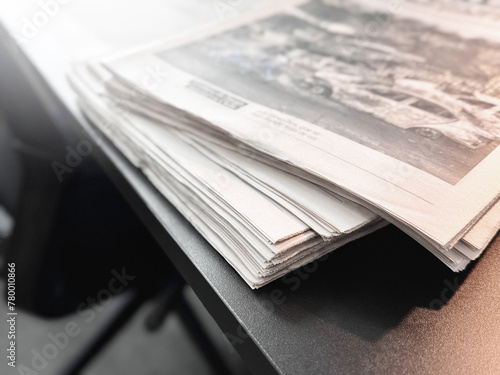 A pile of newspapers in blurred focus is on an office desk illuminated by light. The topic is a printed periodical.