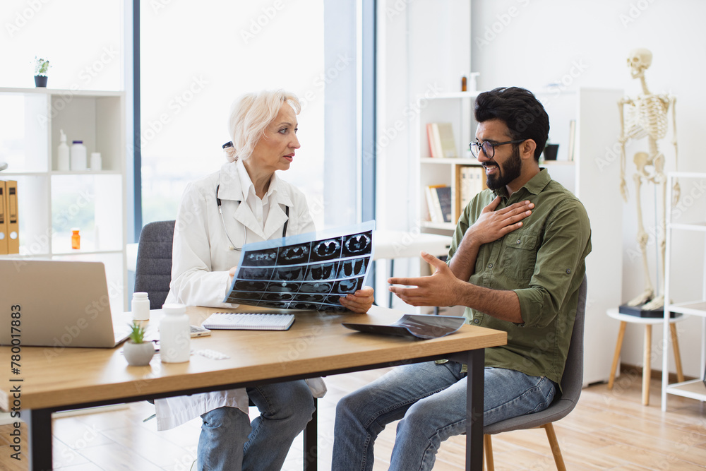 Mindful gray-haired female in lab coat holding MRI scan while brunette guy sitting next to her at desk in hospital. Family doctor examining diagnostic test results while developing treatment plan. Mindful gray-haired female in lab coat holding MRI scan while brunette guy sitting next to her at desk in hospital. Family doctor examining diagnostic test results while developing treatment plan.