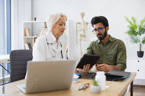 Mature specialist reading out electronic instruction for taking medication correctly. Senior female doctor and male patient sitting at table of modern medical center and watching together at tablet.