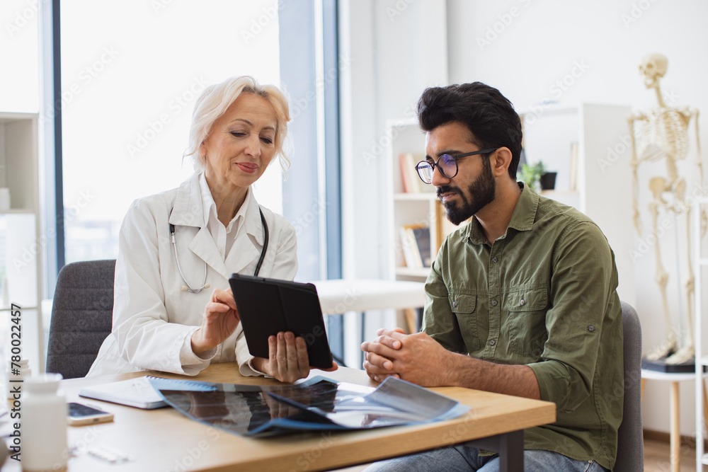 Young gentleman describing symptoms of illness to female doctor. Gray haired physician giving recommendations and using tablet for writing electronic prescription.