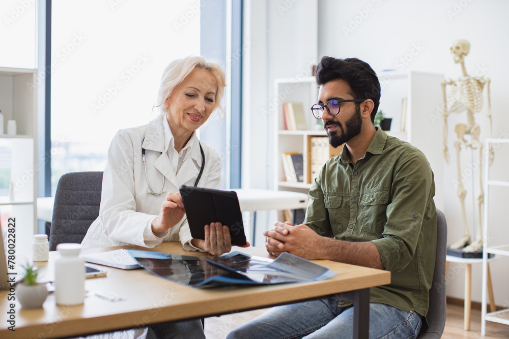 Obraz premium Gray haired physician giving recommendations and using tablet for writing electronic prescription. Young gentleman describing symptoms of illness to female doctor.