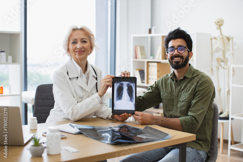 Smiling Senior professional and happy lady posing with CT scan picture on tablet while staying in consulting room. Both medical specialist and patient taking joy in success of treatment therapy.