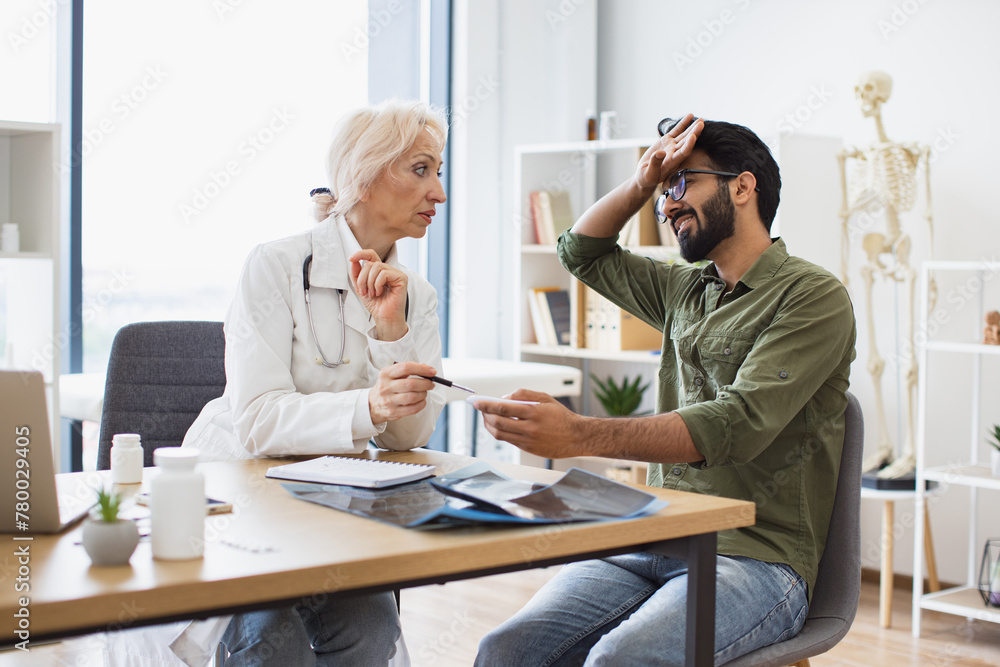 Elder female doctor providing expert check-up and measures temperature to young patient in hospital office. Man with headache listening attentively to recommendations regarding medical history. Elder female doctor providing expert check-up and measures temperature to young patient in hospital office. Man with headache listening attentively to recommendations regarding medical history.