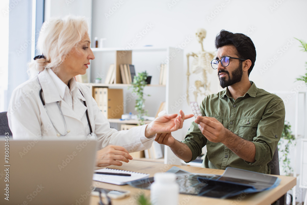 Doctor discussing health issues and reviewing past medical data on laptop. Senior female general practitioner communicating with young patient holding bottle and blister of medication in new clinic.