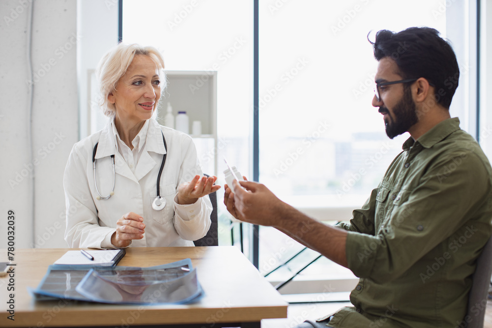 Fototapeta premium Dressed in white uniform senior female therapist consulting young patient at check-up meeting in hospital office. Bearded man keeping bottle and blister of pills and listening to doctor's advice.
