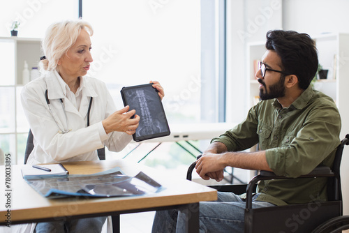 Attractive bearded young man looking on tablet with x-ray image carried by radiologist in consulting room. Confident senior specialist discussing results of diagnostic tests with wheelchair user. photo