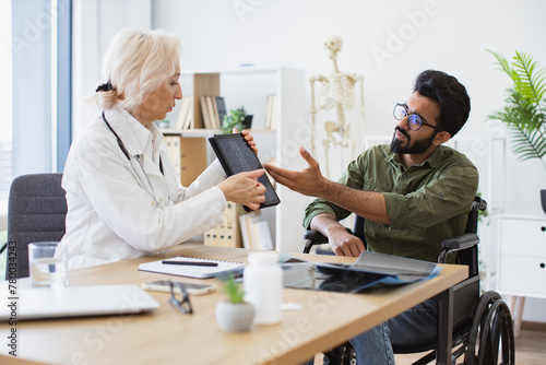 Confident senior specialist discussing results of diagnostic tests with wheelchair user. Attractive bearded young man looking on tablet with x-ray image carried by radiologist in consulting room. photo