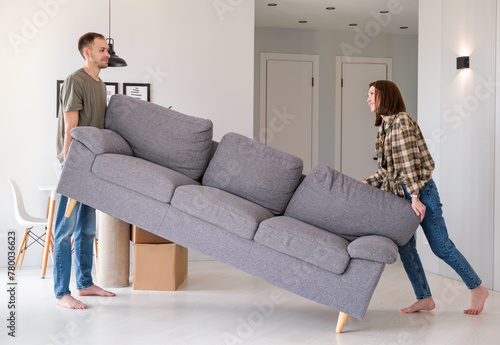 Happy young couple moving sofa in living room, replacing furniture at home, copy space. Millennial husband and wife making rearrangement in their apartment, side view photo