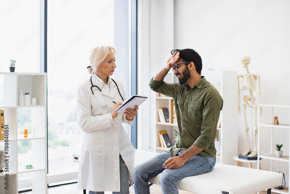 Obraz premium Mature elder physician giving recommendations and using notebook for writing prescription. Bearded gentleman describing symptoms of illness to senior female doctor and gesturing hands.