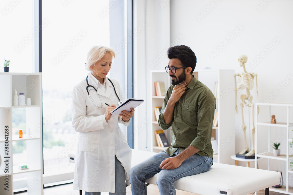 Fototapeta premium Bearded gentleman describing symptoms of illness to senior female doctor and gesturing hands. Mature elder physician giving recommendations and using notebook for writing prescription.