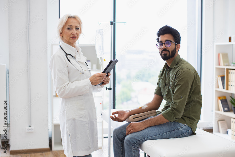 Obraz premium Experienced woman in lab coat using tablet for testing dropper with solution for chemotherapy for young man sitting on couch Healthcare practitioner consulting male patient with cancer in clinic.