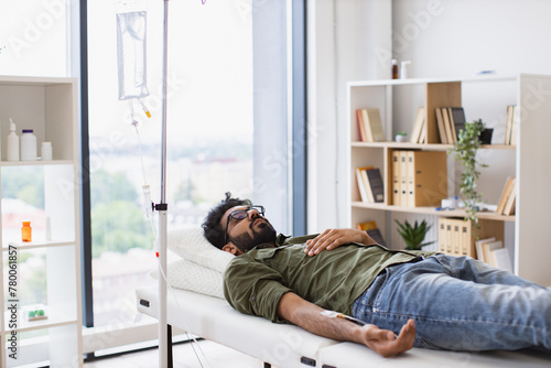 Young bearded man taking dropper with solution for chemotherapy lying on exam couch. Tired sick male in casual wear drips medicine intravenously while lying on modern light ward. © sofiko14
