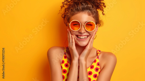 A woman with curly hair and orange sunglasses is smiling and making a silly face. A glad girl wear trendy clothes applause celebrate luck fortune victory isolated on vivid yellow color background