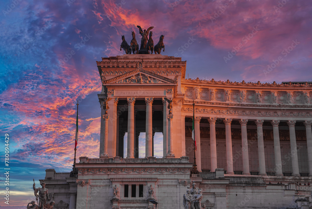 Fototapeta premium Neoclassical building at sunrise or sunset in Italy. Tall columns, pediment with sculptures, and a bronze chariot sculpture on top. Flag with green, white, and red stripes. Latin inscriptions.