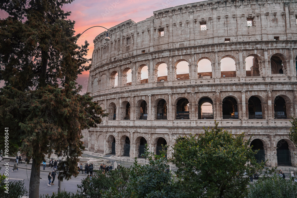 Ancient amphitheater in Rome, Italy, showcasing iconic Colosseum exterior with arches, stone walls, and greenery. Tourists and locals walk near historic site at twilight, capturing serene vibe.