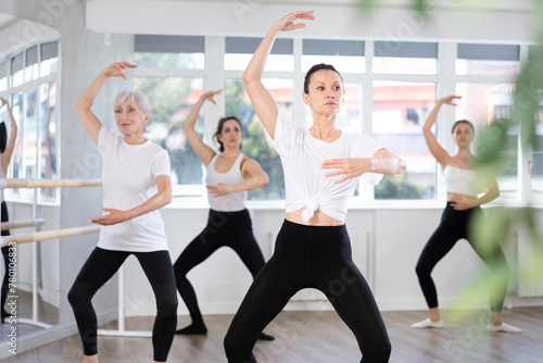 Experienced female ballet dancer, dressed in white top and black leggings, executing dance moves with focused precision among group of younger women rehearsing in studio..