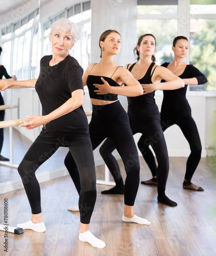 Sporty motivated senior woman participating in barre fitness session with mixed age female group in modern studio, performing graceful demi plie exercise to improve flexibility and strengthen muscles