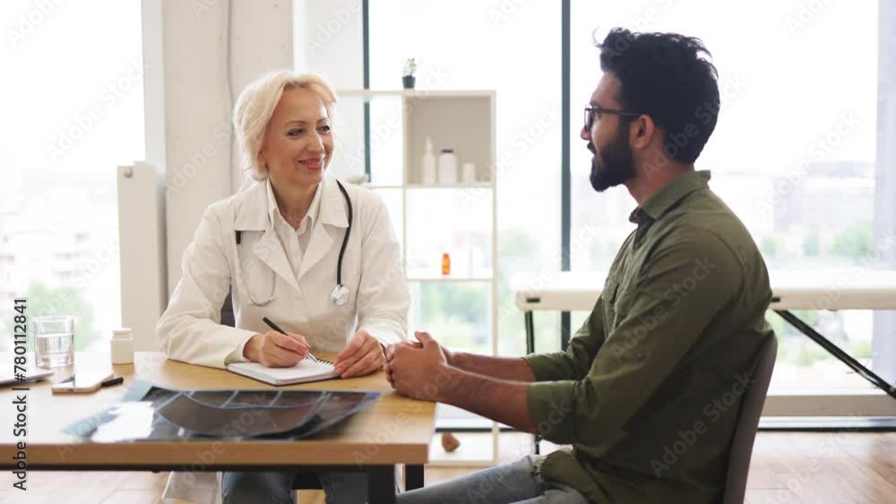 Mature elder physician giving recommendations and using notebook for writing prescription. Bearded gentleman describing symptoms of illness to senior female doctor and gesturing hands. Mature elder physician giving recommendations and using notebook for writing prescription. Bearded gentleman describing symptoms of illness to senior female doctor and gesturing hands.