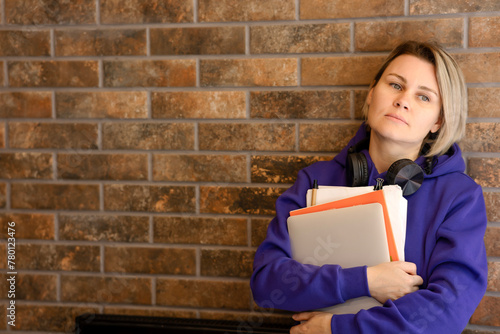 The student girl is thoughtful, looking away, leaning against a brick wall. Young blonde woman in blue hooded sweater holding laptop and notebooks, big black headphones, space for copy