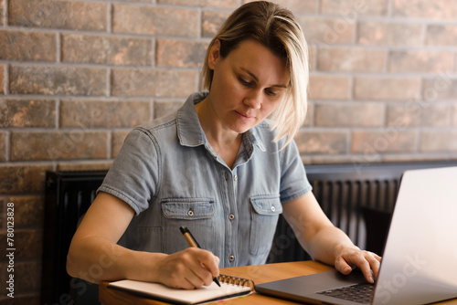 Young blonde woman in blue dress writing notes in notebook, laptop on table. Girl working in office, on wooden table in laptop office, freelancing, business woman