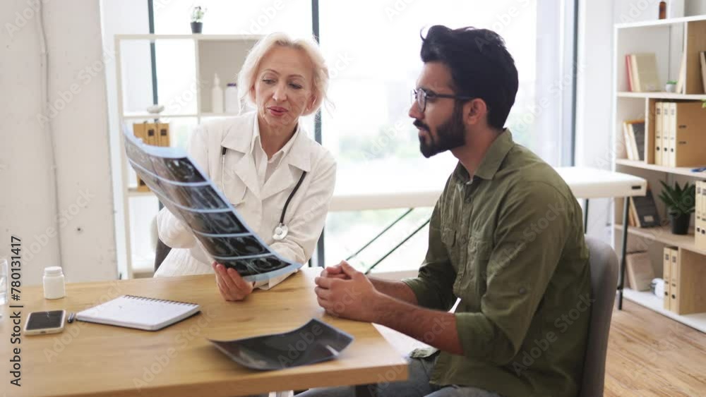 Family doctor examining diagnostic test results while developing treatment plan. Mindful gray-haired female in lab coat holding MRI scan while brunette guy sitting next to her at desk in hospital. Family doctor examining diagnostic test results while developing treatment plan. Mindful gray-haired female in lab coat holding MRI scan while brunette guy sitting next to her at desk in hospital.