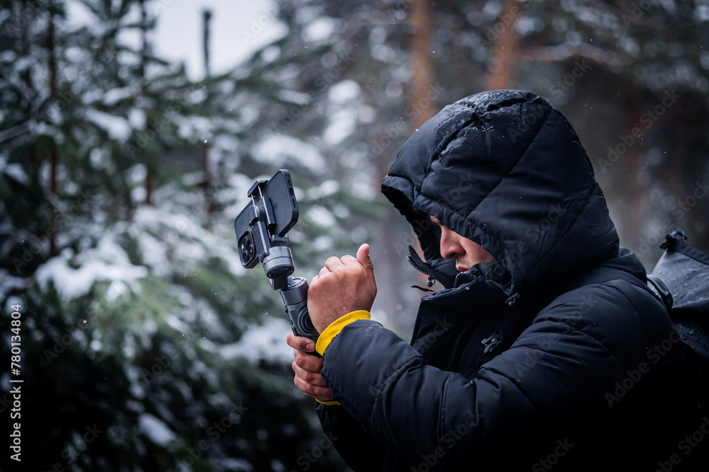 Profile photo of young man in black coat and hood filming with mobile phone and gimbal.