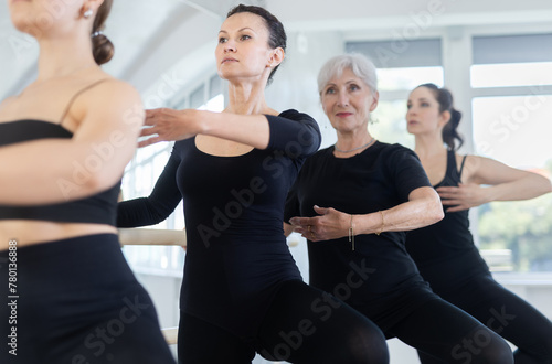 Focused graceful woman mastering passe ballet move at barre during rehearsal with group of dancers of different ages in modern choreography studio