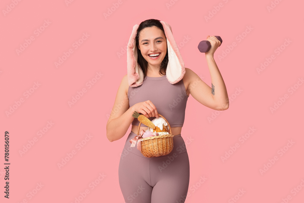 Happy female fitness coach in bunny ears with Easter basket and dumbbell on pink background Happy female fitness coach in bunny ears with Easter basket and dumbbell on pink background