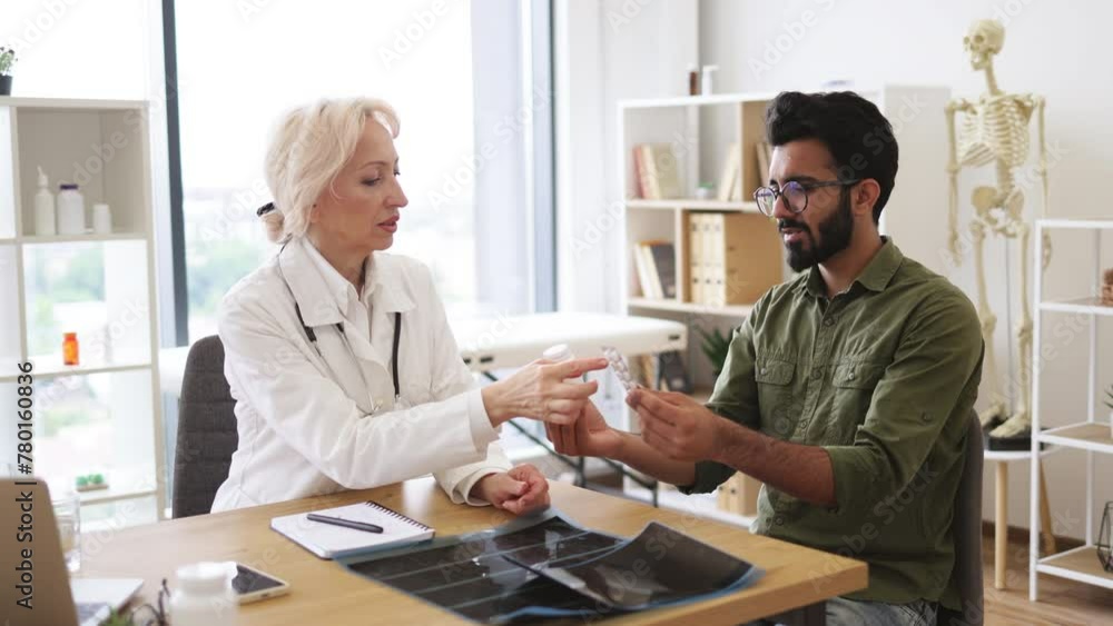 Doctor discussing health issues and reviewing past medical data on laptop. Senior female general practitioner communicating with young patient holding bottle and blister of medication in new clinic. Doctor discussing health issues and reviewing past medical data on laptop. Senior female general practitioner communicating with young patient holding bottle and blister of medication in new clinic.