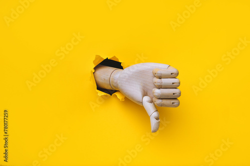 A wooden hand protrudes from a torn hole in yellow paper and shows a thumbs down (dislike). The concept of disapproval and that things will go badly. Artificial intelligence, robot.