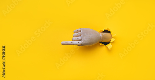 A wooden hand protrudes from a torn hole in yellow paper and points its index finger to the left. Concept of direction, adjustment and assistance. Mannequin, robot photo