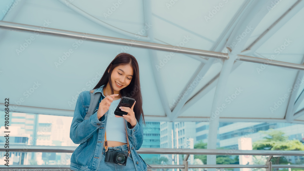 Young asian woman smiling using mobile smart phone outdoor. Happy female tourist wearing jeans jacket and holding smartphone at public - obrazy, fototapety, plakaty 