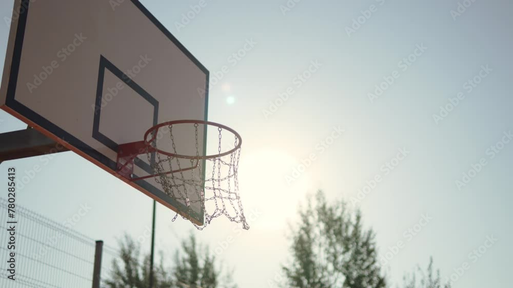 Basketball ball hits hoop in sunlight and causes chains to swing, arc shot. Hit target and earn points on street basketball court. Playing sports and achieving goal in competition outdoor. Basketball ball hits hoop in sunlight and causes chains to swing, arc shot. Hit target and earn points on street basketball court. Playing sports and achieving goal in competition outdoor.