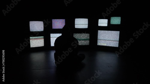 An anxious and isolated person sits in front of a wall of white noise filled televisions media screens in a dark room.