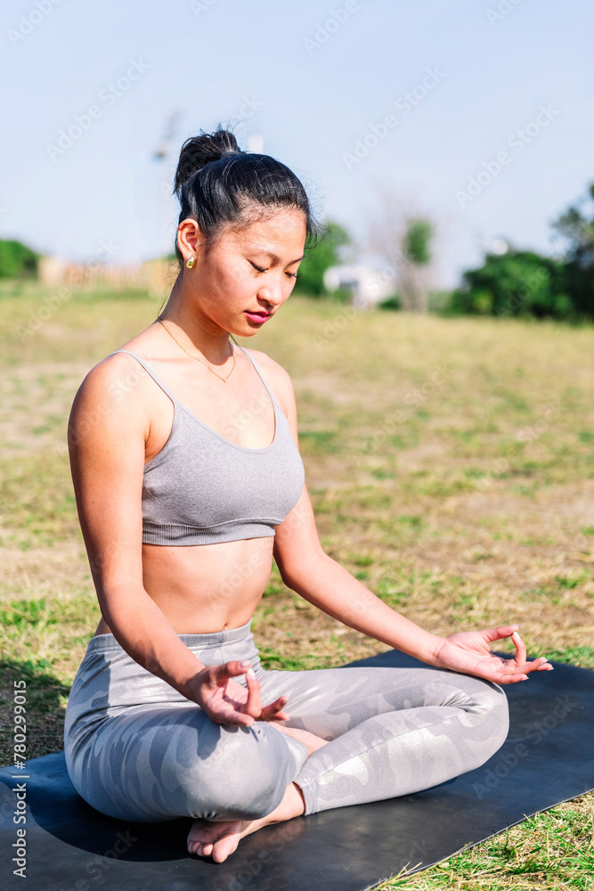 young asian woman doing meditation at park sitting on a yoga mat, concept of mental relaxation and healthy lifestyle