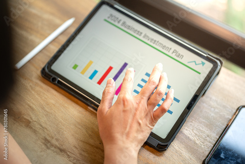 Action of a person hand is touching on tablet screen to review the 2024 business investment plan with graph and chart data. Business working with technologyo concept, close-up and selective focus.
