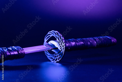 Close-up of samurai sword replica with a decorative hilt, illuminated by a neon light. Intricate details of the sword are highlighted against the colorful backdrop.