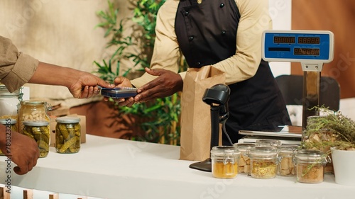 Vegan customer using card at pos to pay for natural food alternatives, purchasing homemade sauces and pantry supplies. African american man shopping for organic additives free products at local market