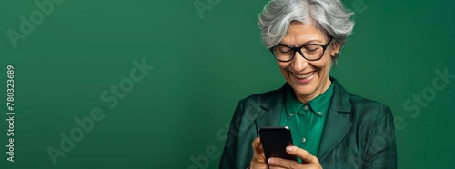 Photo of A mature businesswoman with short silver grey hair and glasses using her smartphone on green background, copy space for text. Web banner showing a woman smiling while looking at phone