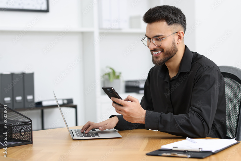 Handsome young man using smartphone while working with laptop at wooden table in office Handsome young man using smartphone while working with laptop at wooden table in office