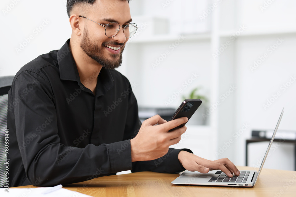 Handsome young man using smartphone while working with laptop at wooden table in office Handsome young man using smartphone while working with laptop at wooden table in office
