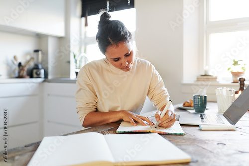 A woman sits focused in her kitchen and sketching a design in a book. On the table is an open laptop and there are several open notebooks photo