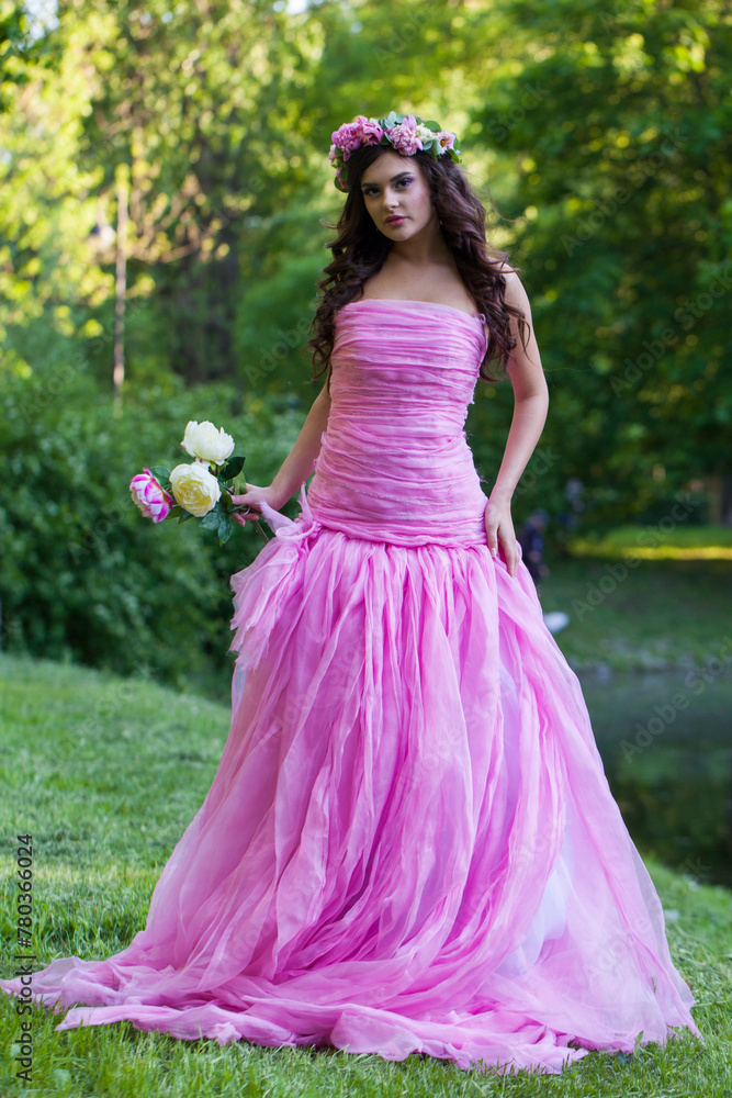 Romantic female model with bright makeup, curly hair and pink flower wreath on green summer foliage background. Healthy woman in pink dress portrait Romantic female model with bright makeup, curly hair and pink flower wreath on green summer foliage background. Healthy woman in pink dress portrait