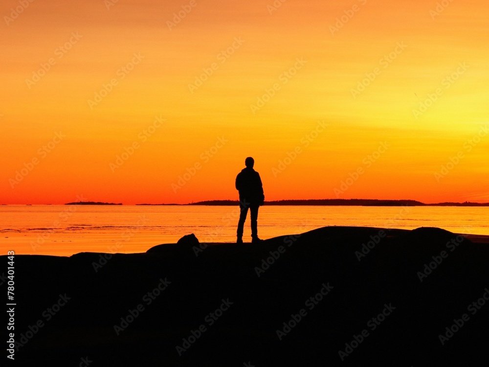 man standing on beach looking out to sea at sunset, from rocks in foreground